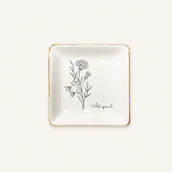 Personalized square gold rim ceramic ring dish with birth flower line art on a cream background